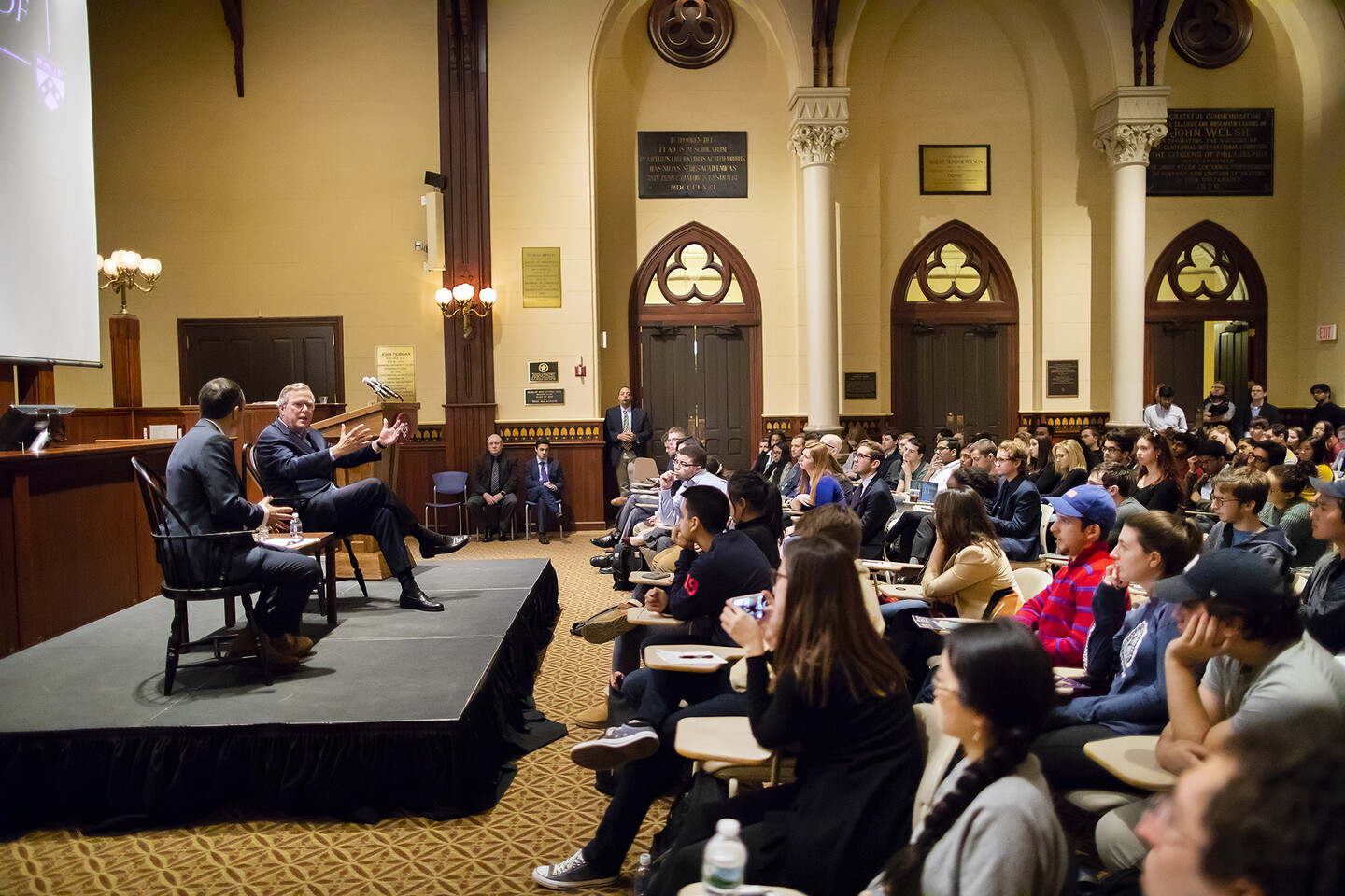 bush-and-green-seated-on-stage-facing-audience-of-students
