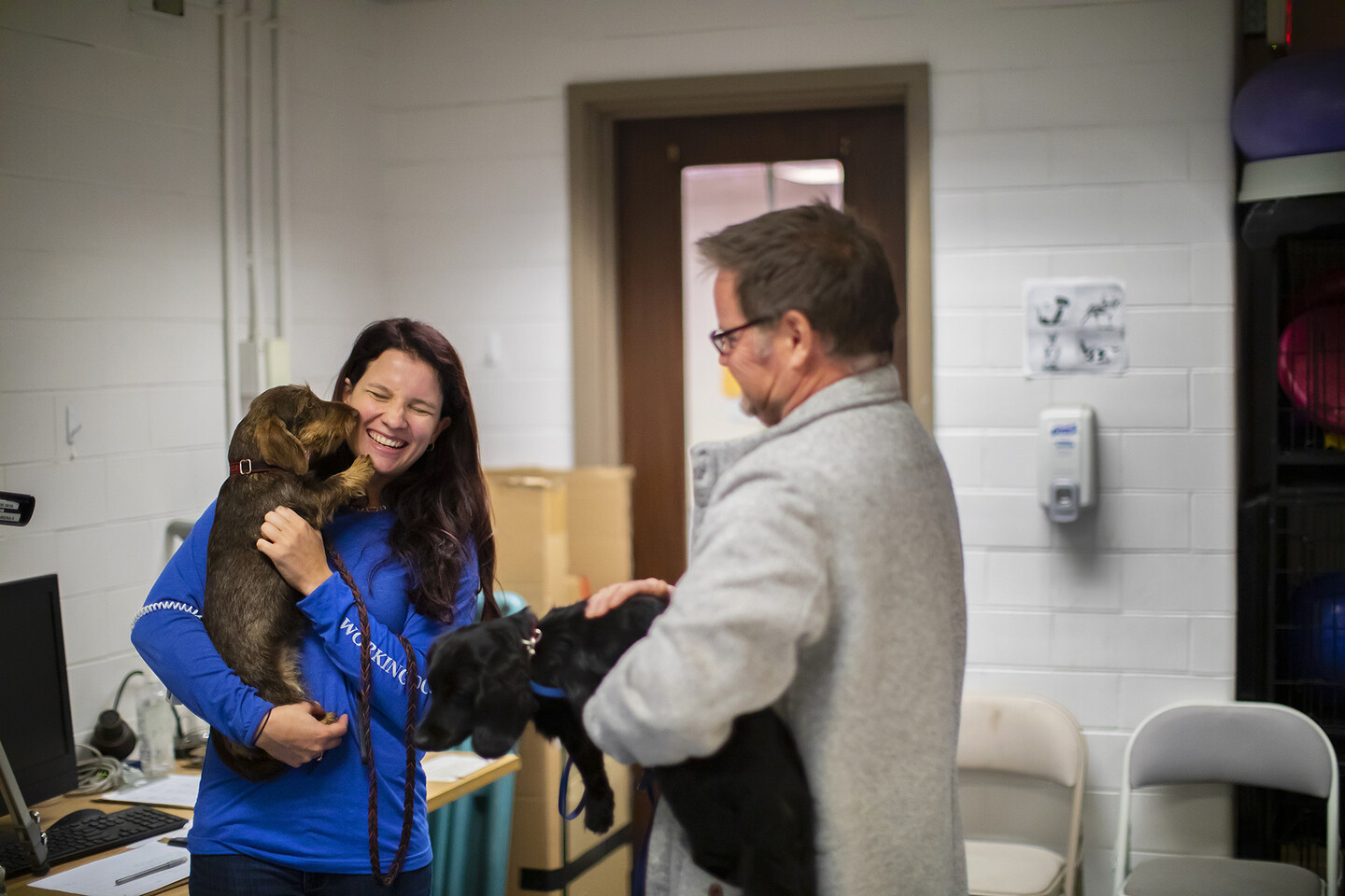 Course instructor Meghan Ramos holds a dachsund and smiles as it licks her cheek as a course participant, also holding a dachsund, looks on