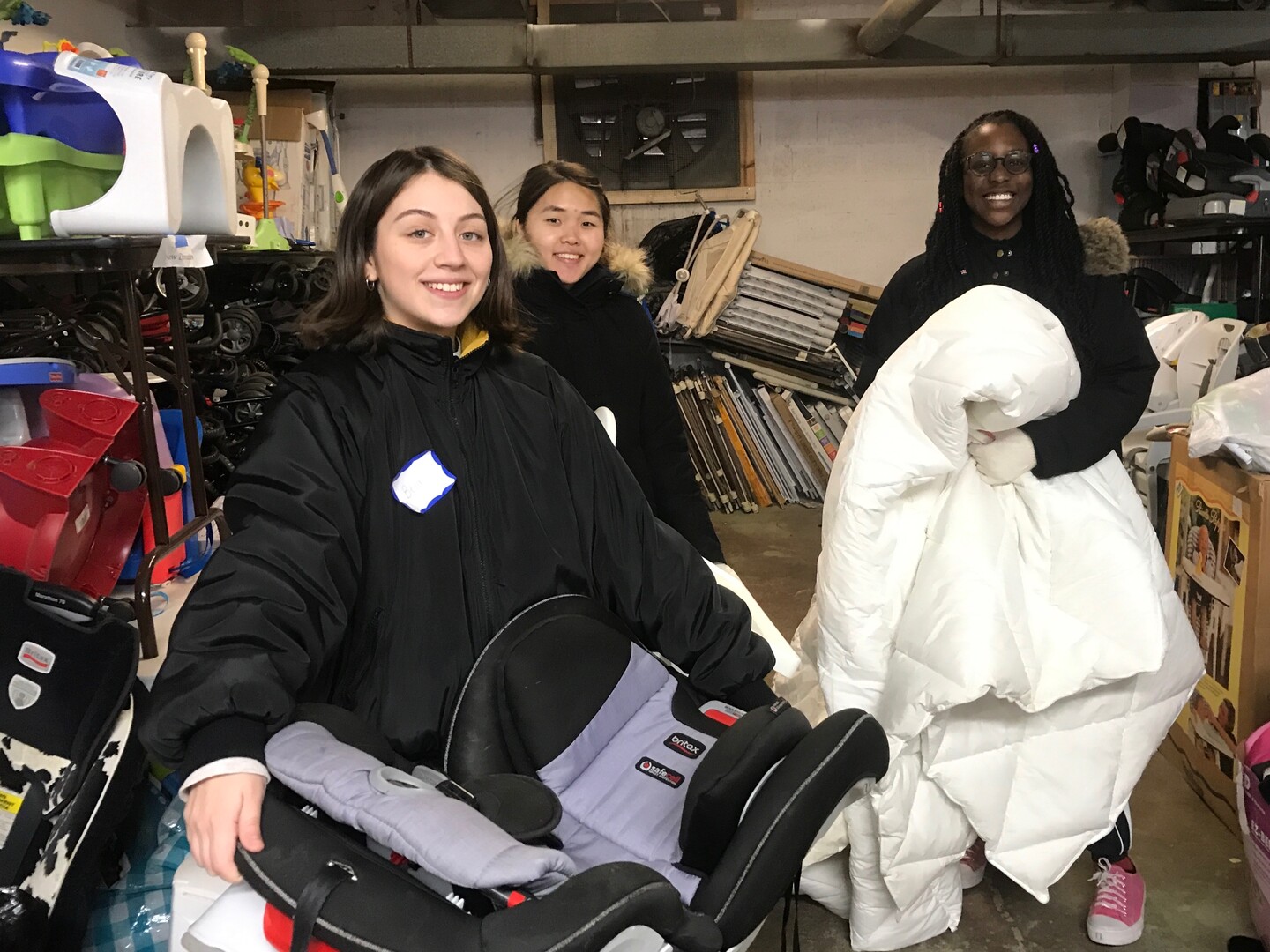 Three students standing in warehouse filled with home items, one carrying a blanket and the other a child's car seat. 