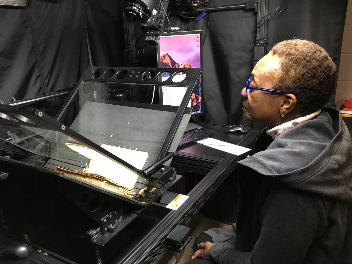 Woman operating equipment to digitize a book with book open on glass surface next to a computer displaying the image.