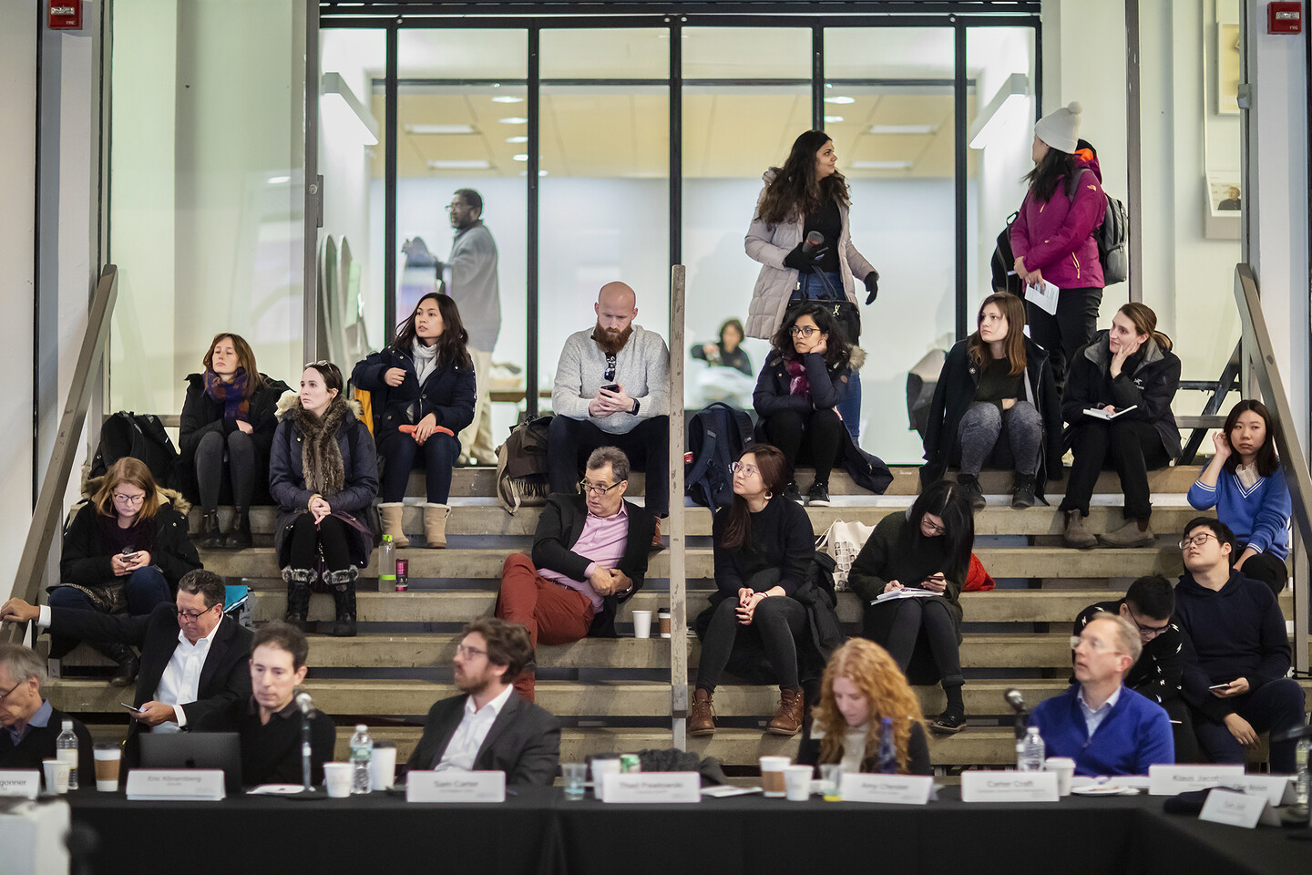 Audience members sit at a table and, beyond, on stairs, paying attention to a presentation.