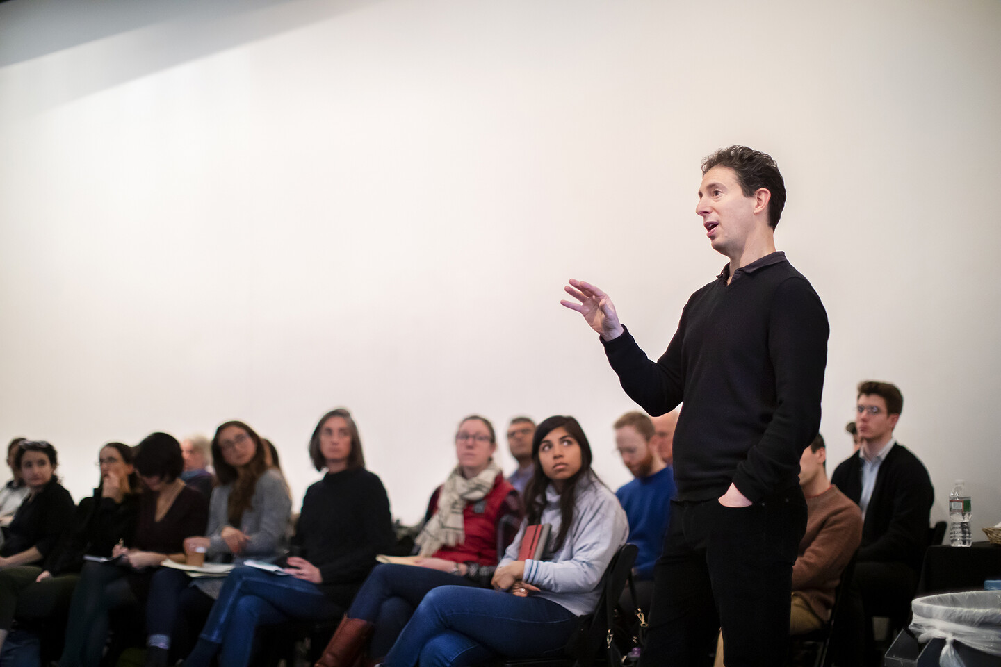 A man stands, hand gesturing in the air, as others, seated, look on.