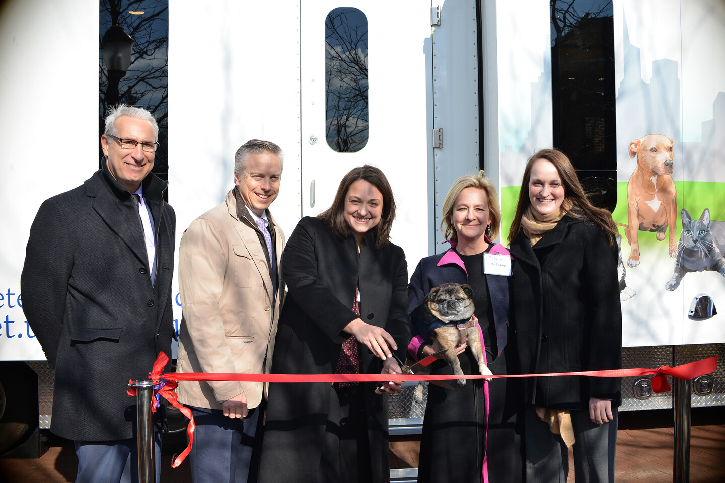 Five people stand in front of a trailer with dogs and cats featured on it, cutting a red ribbon and smiling.