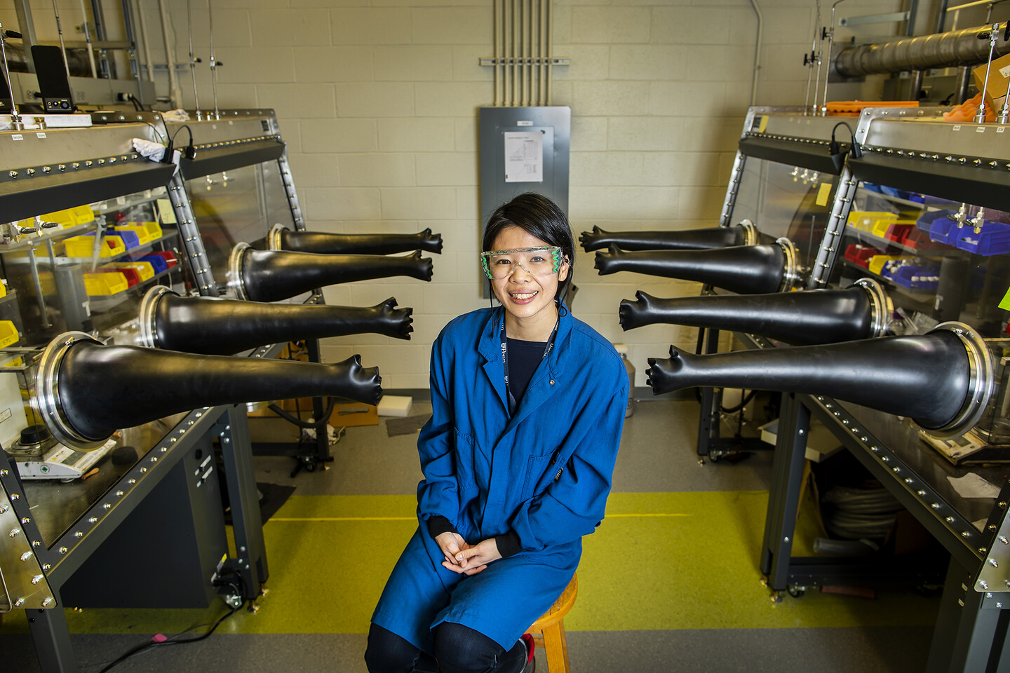 chu seated in a lab with rubber gloves outstretched from glass-covered laboratory benches
