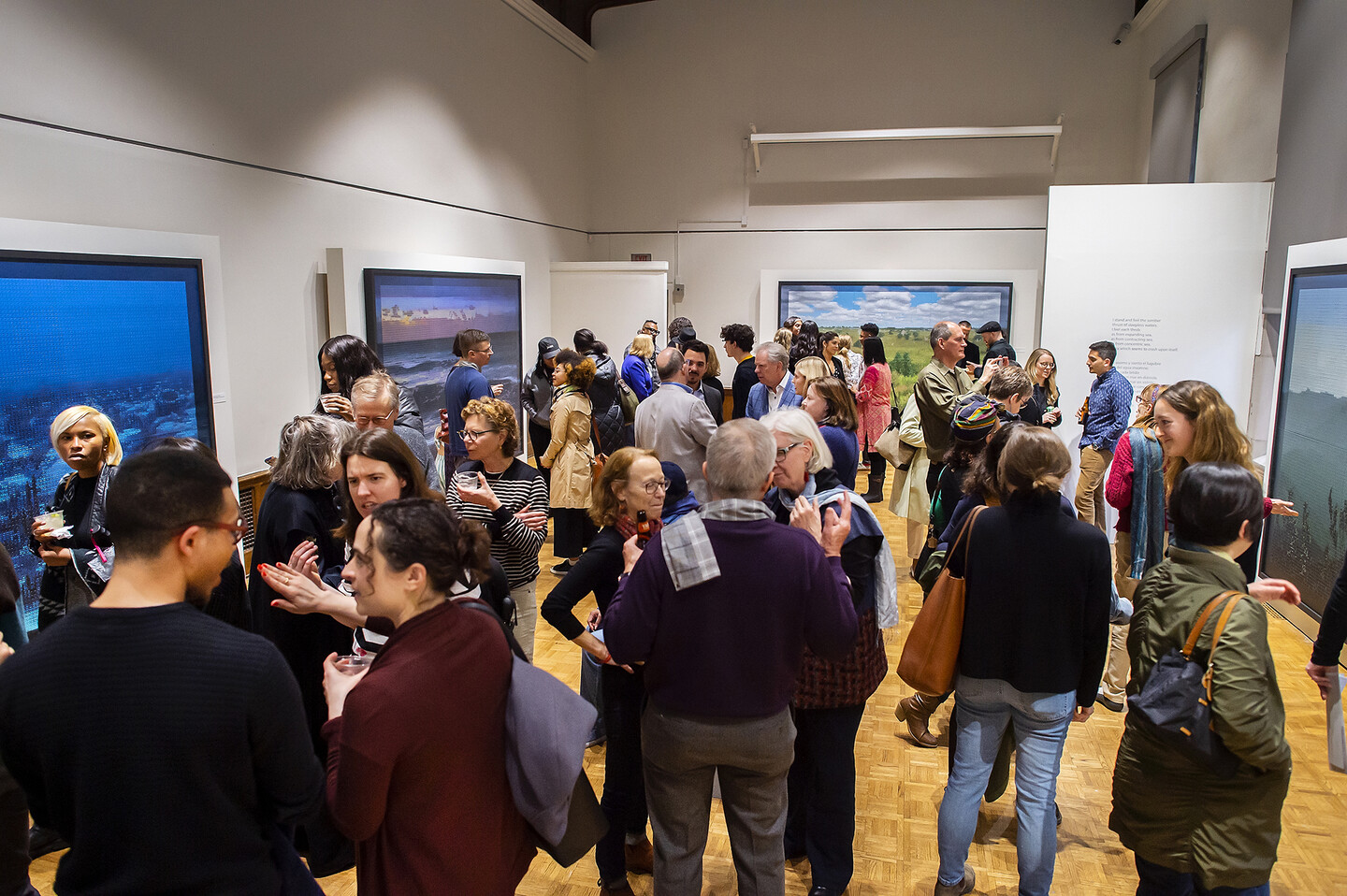 Many people in the art gallery with the paintings visible around the perimeter. 