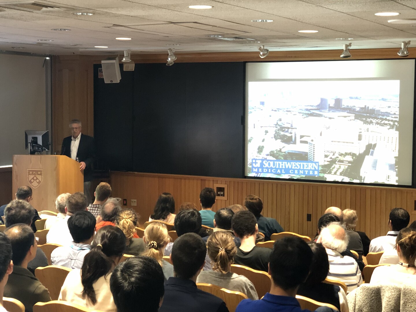 a professors standing at the front of a lecture hall giving a powerpoint presentation