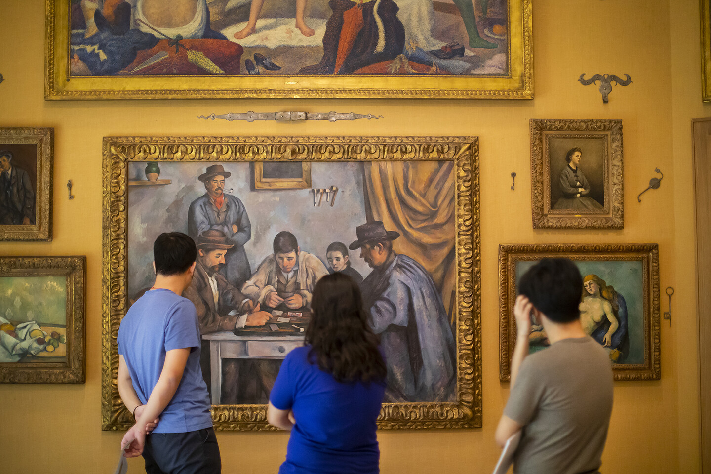 Three people with their backs to the camera standing in front of several painting hung on a yellow wall.