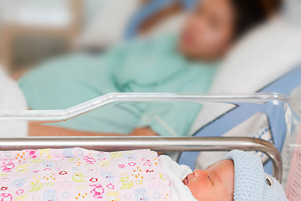 newborn baby in bassinet beside hospital bed with birth mother in background