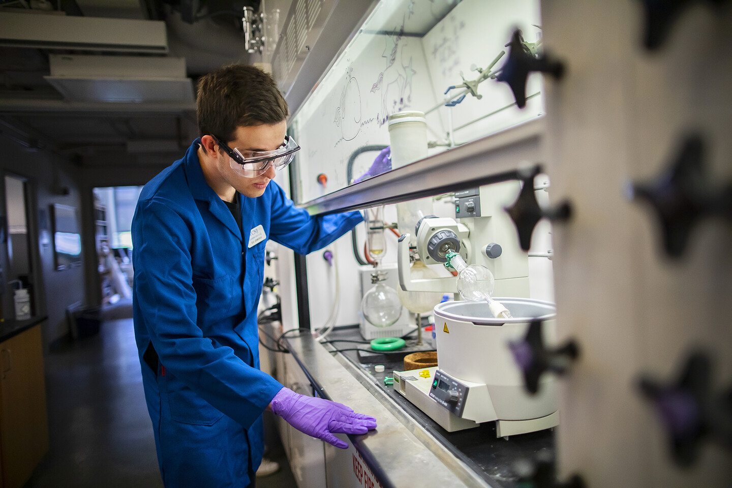 a person standing next to a chemistry lab hood looking at a flask that is held in place by a mechanical device