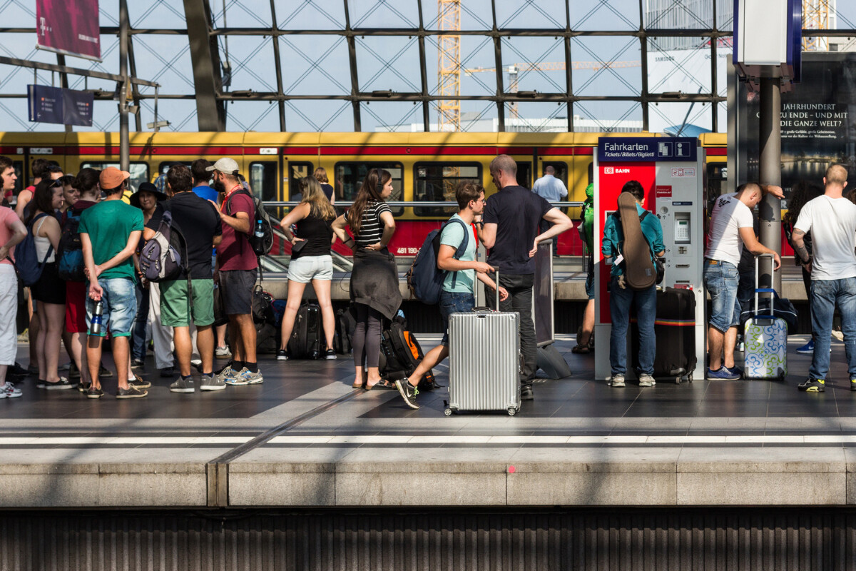 A group of people waiting on a platform of a train station with sunlit windows and a train on the tracks.