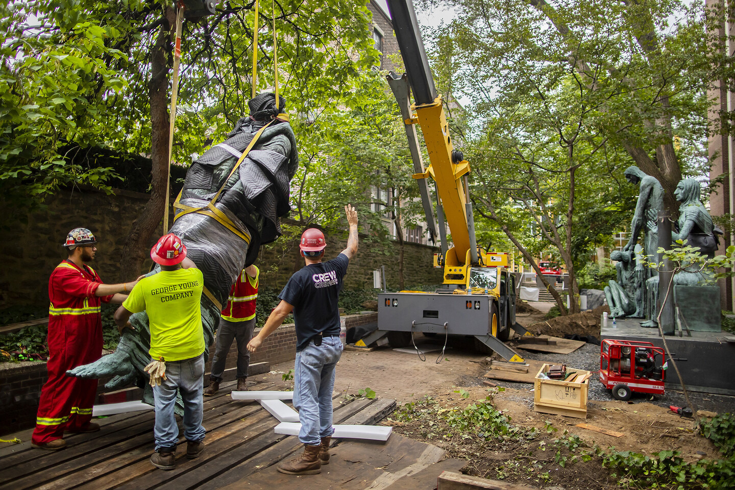 Large sculpture figure suspended by a large crane with construction workers guiding it in a garden setting with a brick wall behind them. 