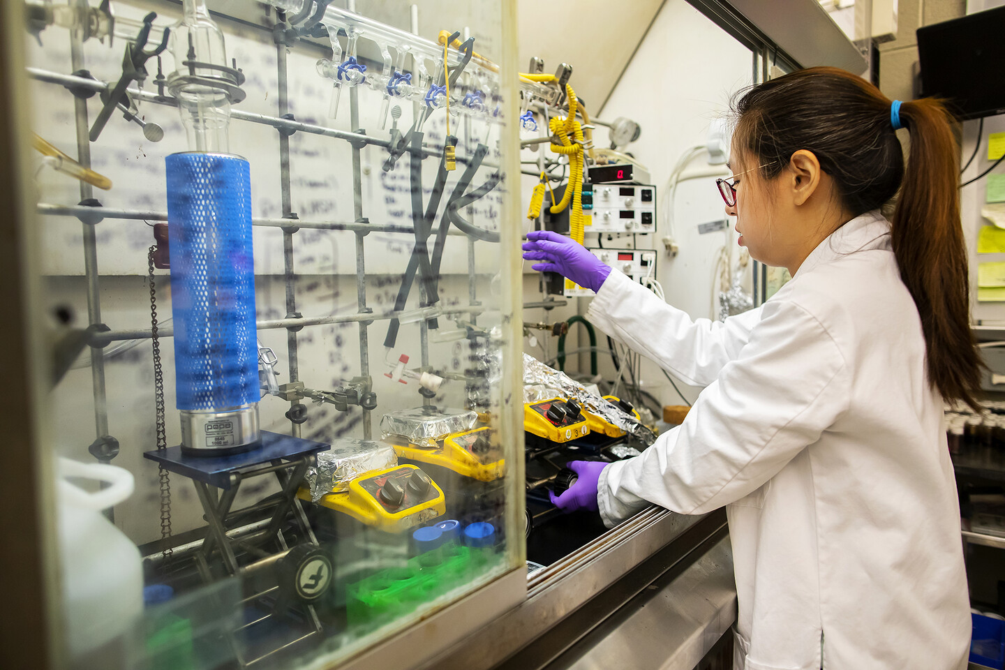 lee reaching for a test tube while working in a chemistry hood, the glass is covered in chemical equations and the hood is full of glassware, tubing, and hot plates