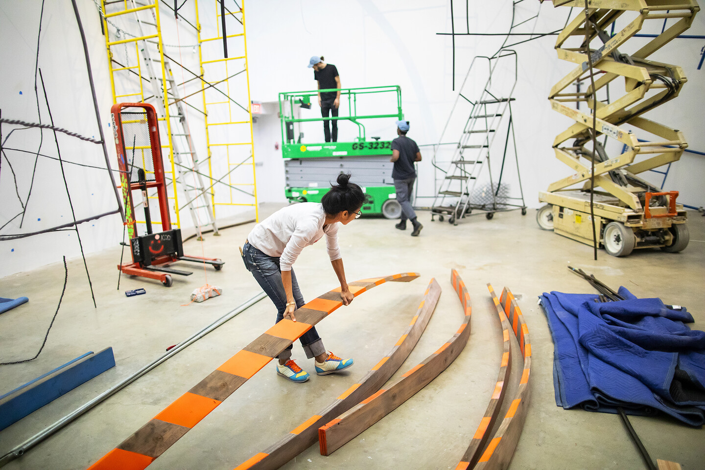 Artist picking up long wooden plank in gallery during sculpture installation. 