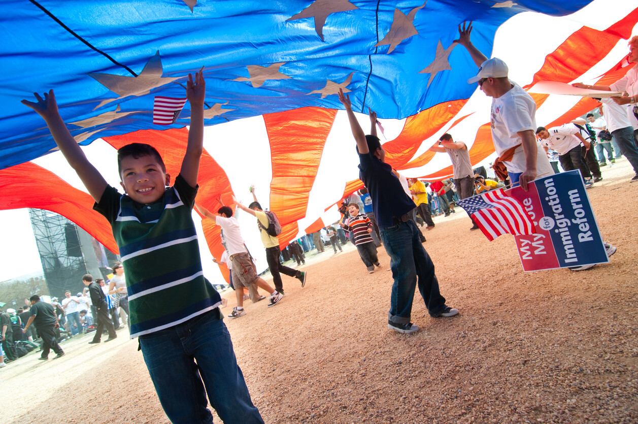 Several adolescents stand under a giant American flag like a parachute aloft at an immigration reform rally.