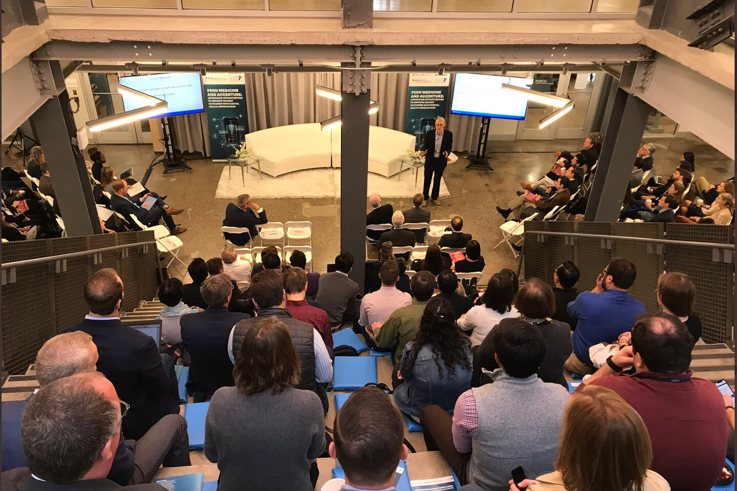 a person standing in front of a large room of people the viewpoint from the back of a tall set of bleachers in an industrial room