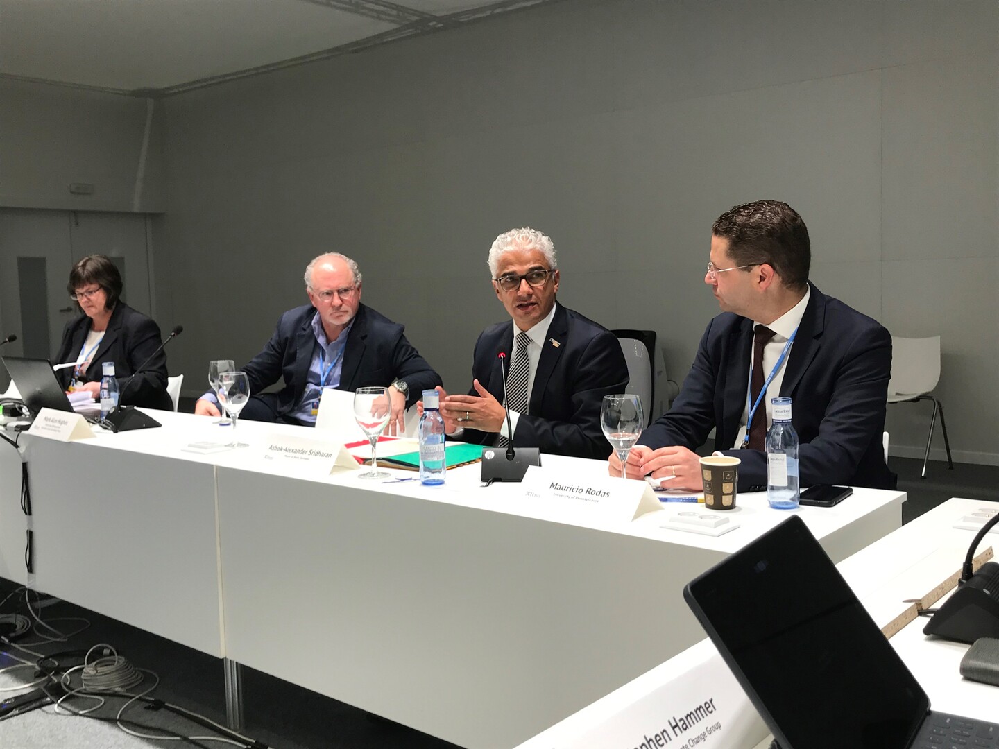 Four people sitting at a white table, engaged in discussion. They have nameplates in front of them. The two on the right are visible, reading "Ashok-Alexander Sridharan" and "Mauricio Rodas"