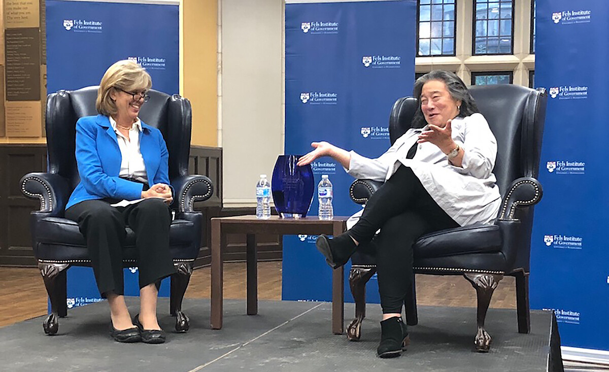 Two people sit in wing-backed armchairs, separated by a small side table with an empty vase and two plastic water bottles, smiling and having a conversation in front of a wall with the words Fels Institute of Government and the Penn logo printed on it.