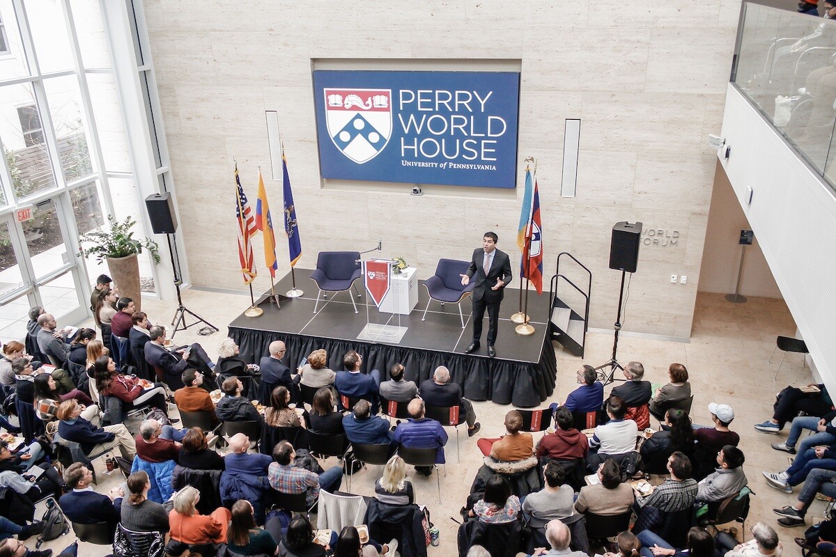 A person on a stage speaks to a crowd of people sitting in chairs, with a sign reading Perry World House behind him.