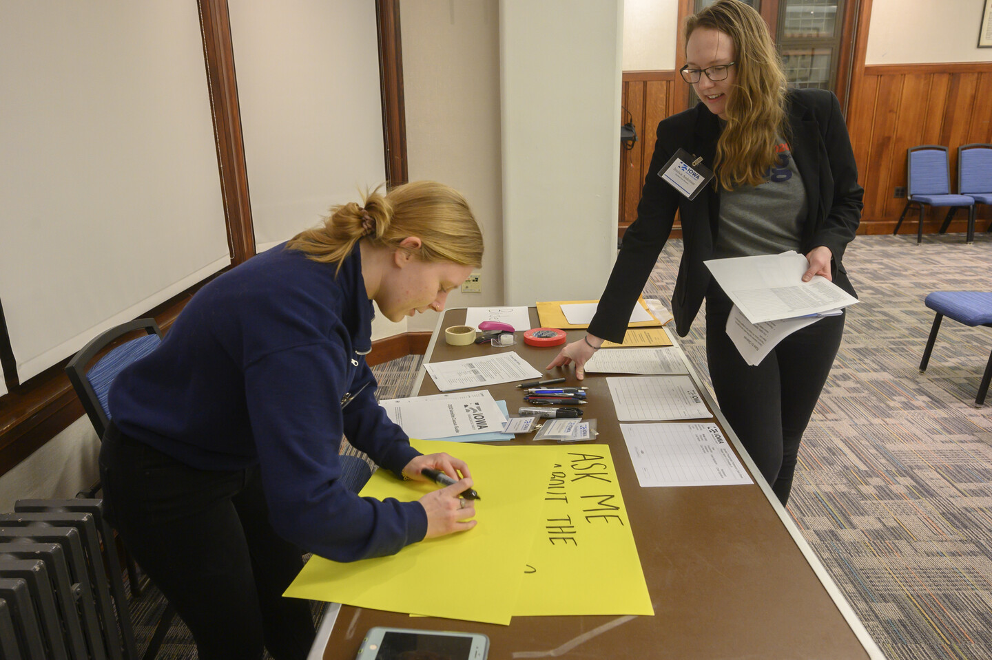 A person writing on a large yellow posterboard at a brown table, with another person looking on.