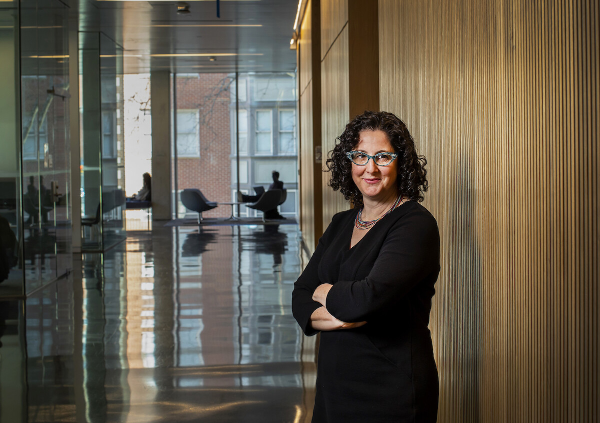 A person in a black dress and glasses stands against a wooden wall with arms crossed, looking at the camera, in the background is a huge window and people in chairs in front of the window.