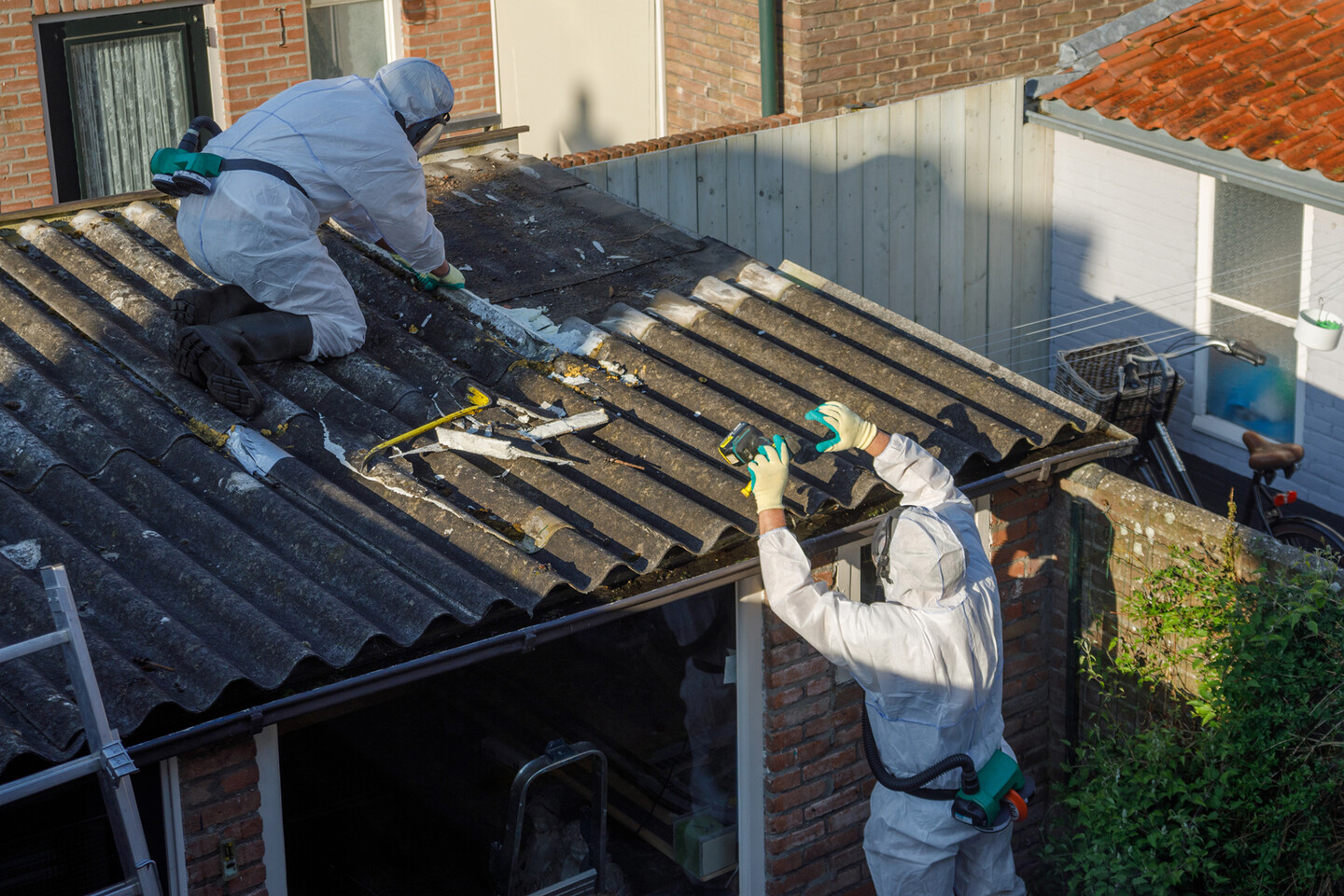 One worker on the roof of a one-story building and one worker on the ground working together and wearing boots, gloves, masks and protective clothing.  