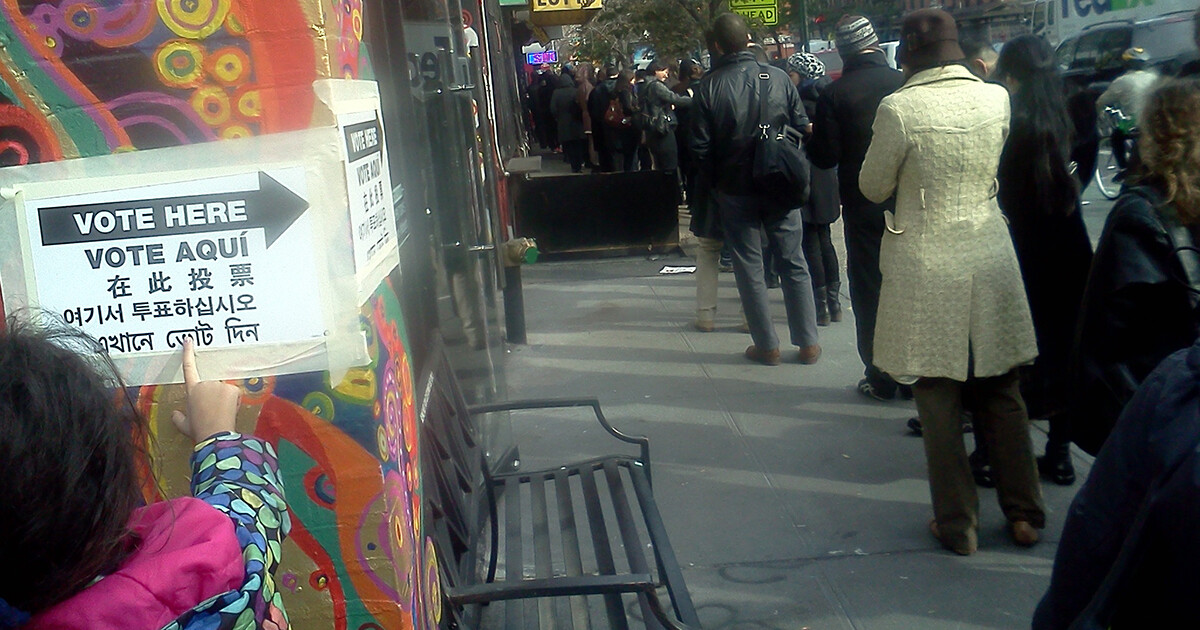 A line of people waiting to vote outside on a city sidewalk, a child points to a Vote Here sign taped to a colorful wall painted with a mural.