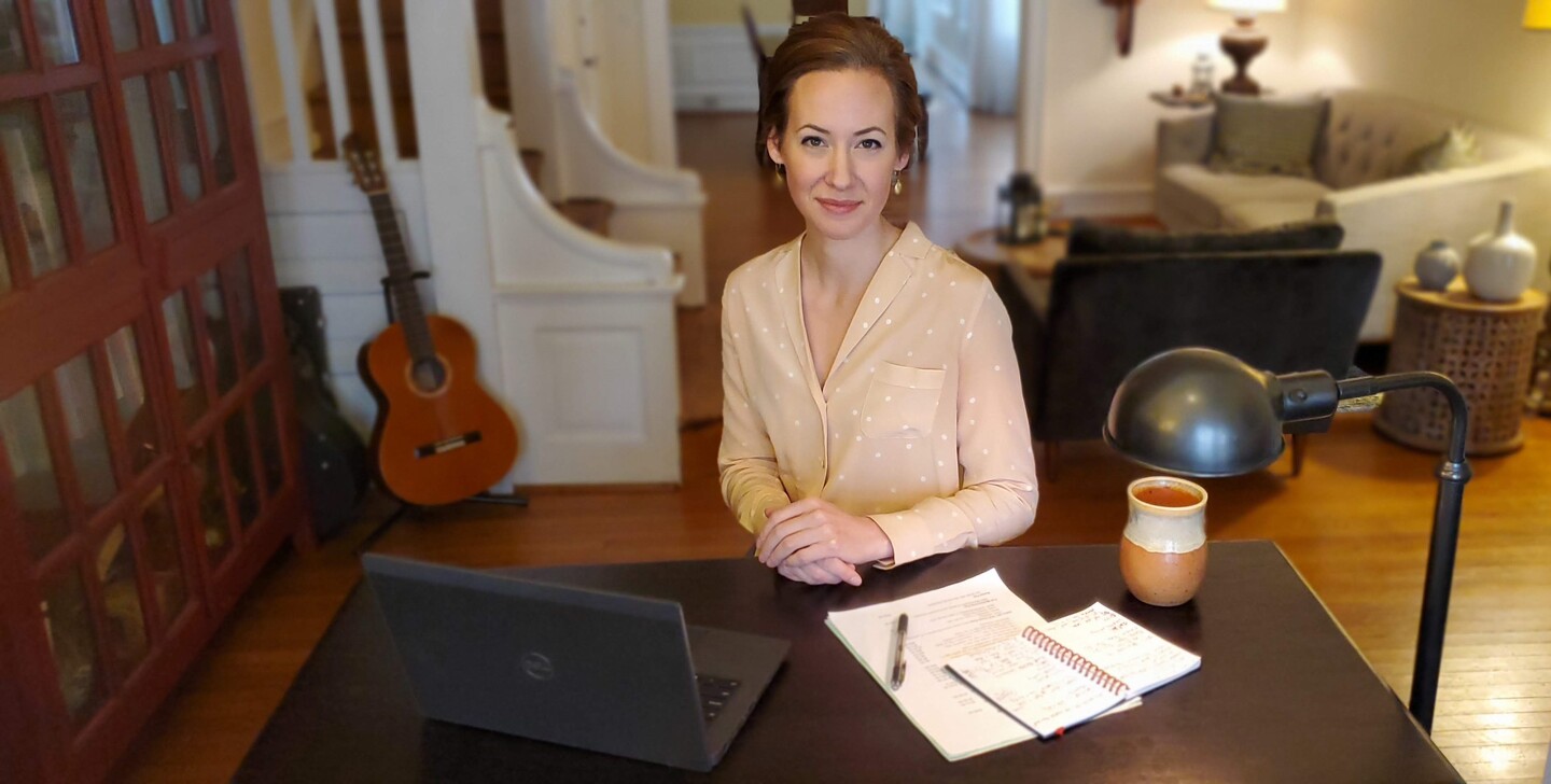 Librarian sitting at a desk in her home with a laptop computer, a notebook, a lamp, and a mug on the desk in front of her.