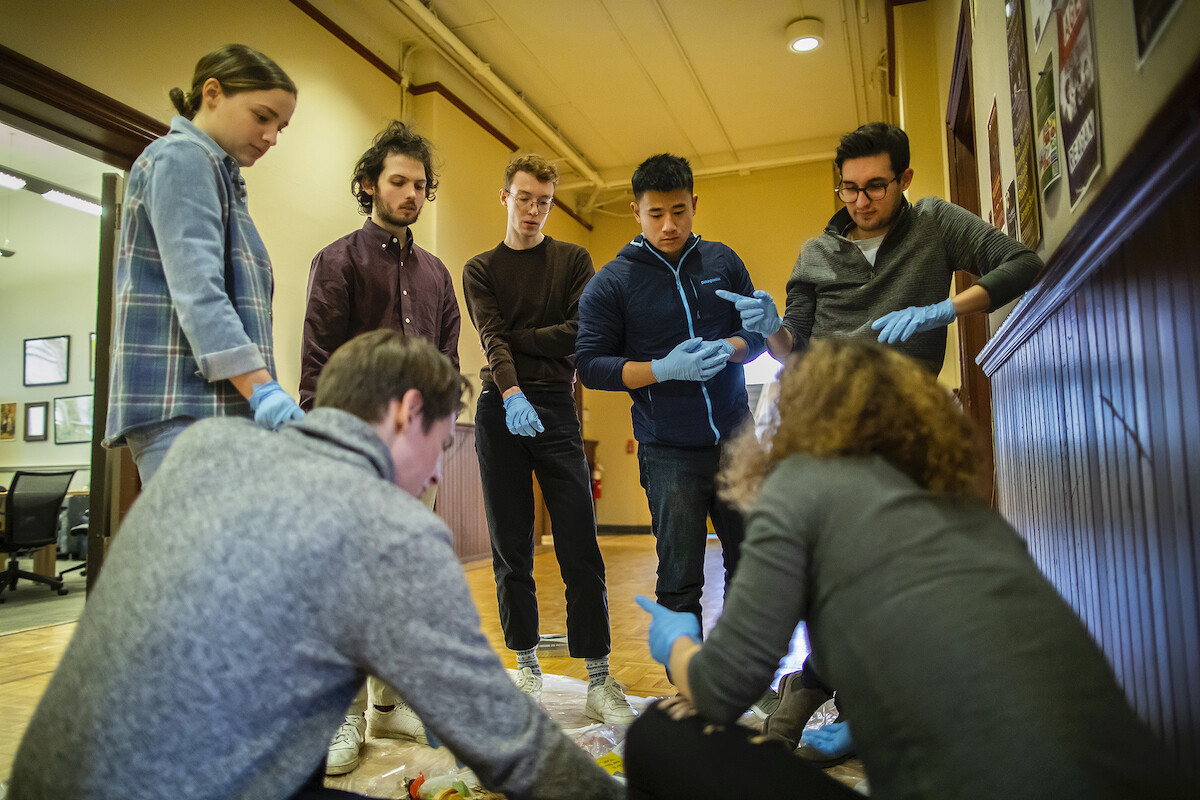 Four people wearing latex gloves look down at a pile of trash in a hallway, while two other people squat beside it, backs facing the camera