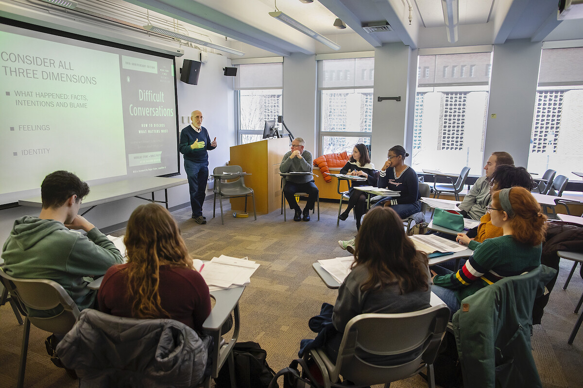 A group of students sits at desks arranged in a semi-circle as an instructor stands by a screen displaying the words "difficult conversations"