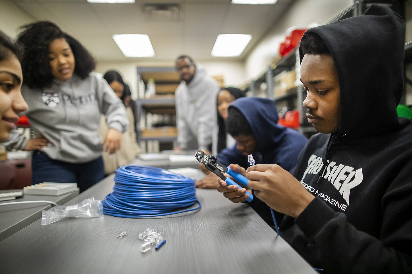 Young man in hoodie uses pliers on coated wire as others look on