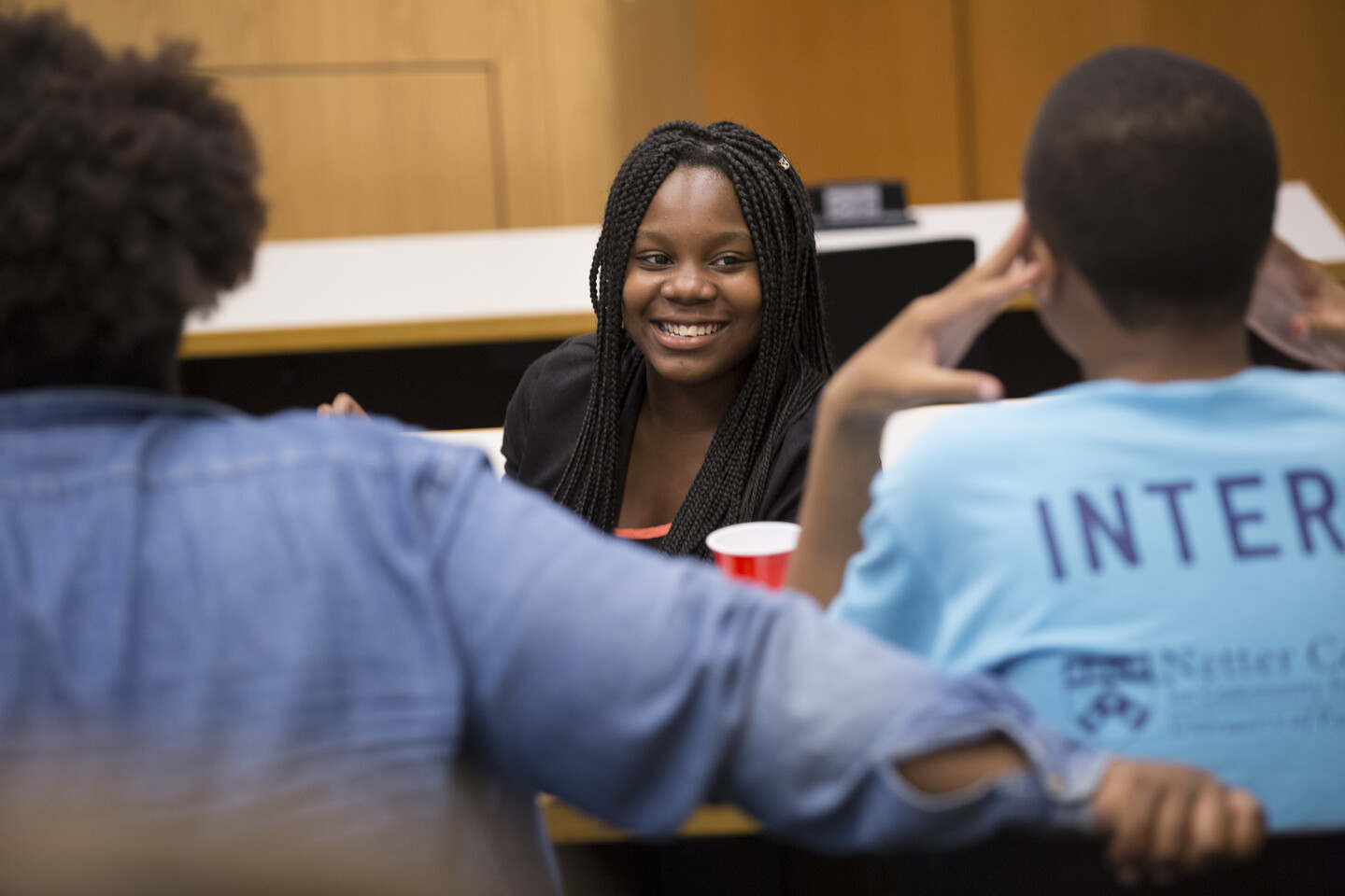 a smiling young woman with braids is pictured; two other students have their backs to the camera