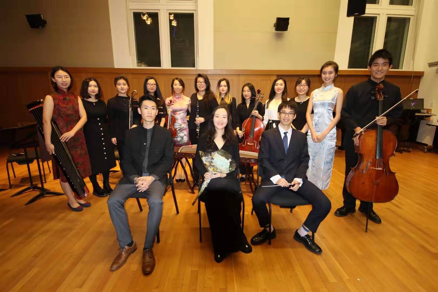 Three people seated and eleven people standing behind them holding various instruments in a practice room. 