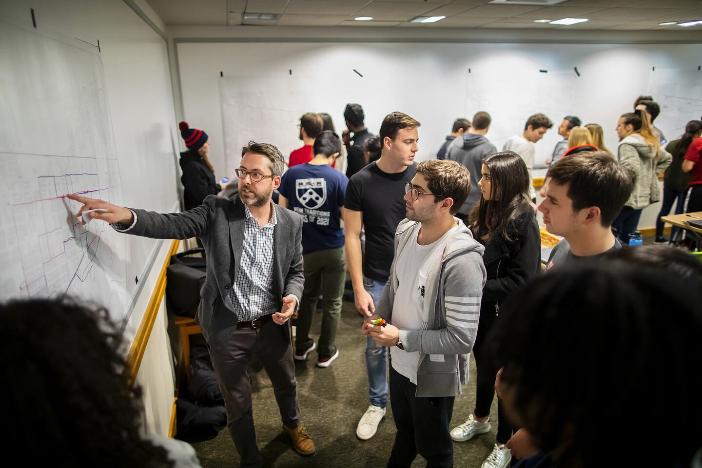 Brent Cebul points to a large map on a wall surrounded by students standing nearby in a classroom. 