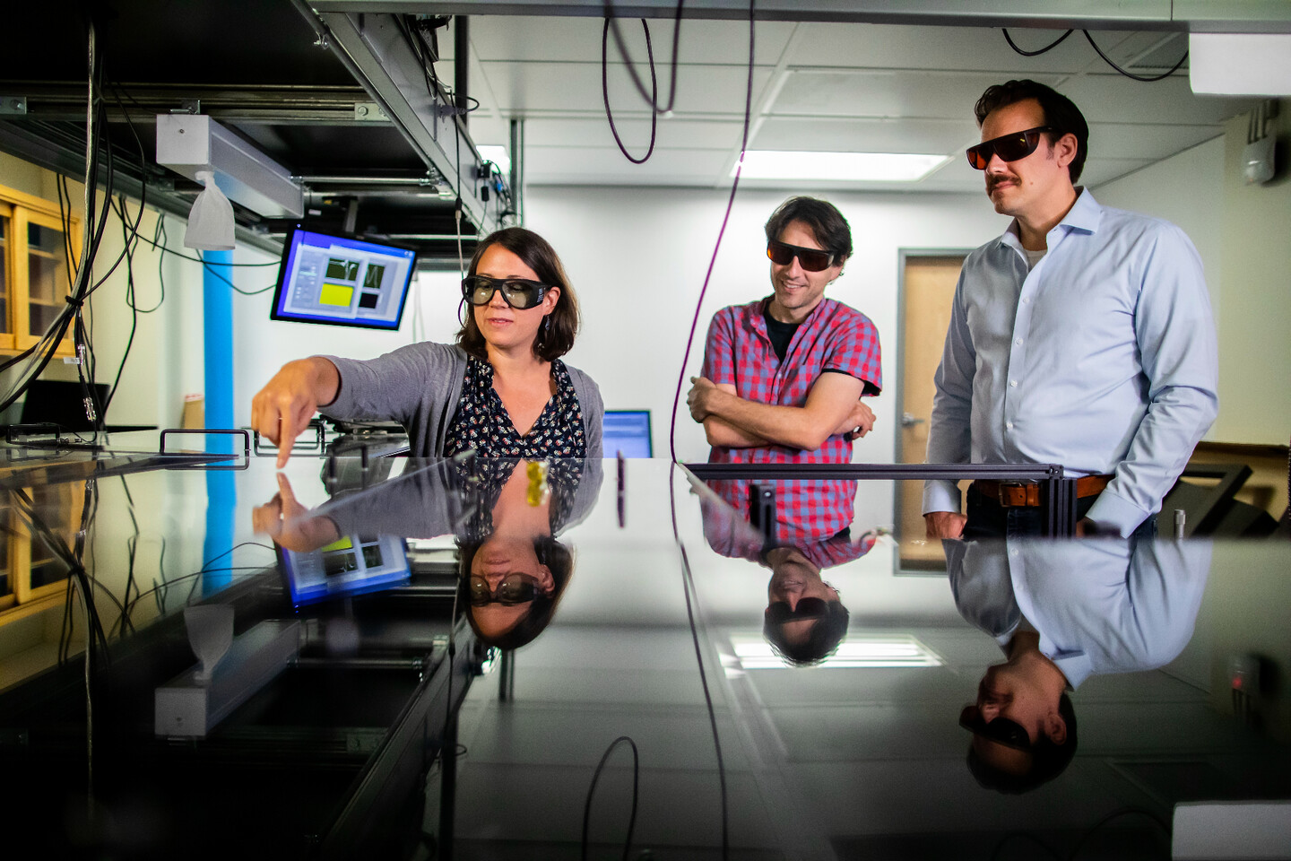 three people looking down at an optics table with computer screens and wires in the background