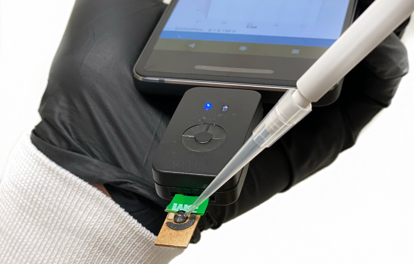 A hand in a black rubber gloves holding a cell phone with a sensor attached. A dropper is touching the sensor.