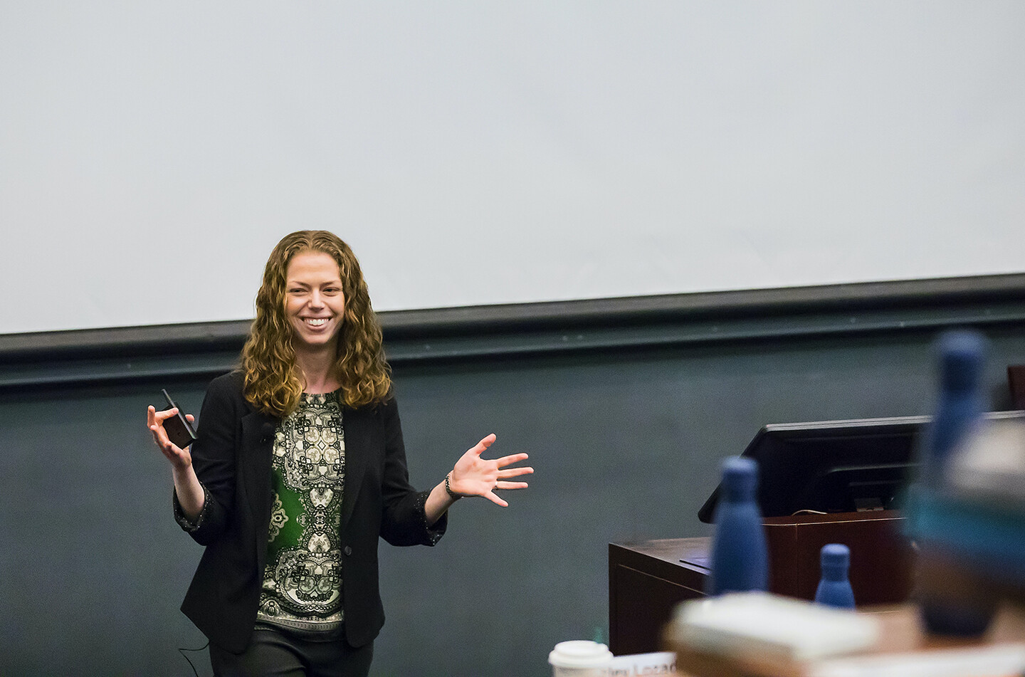A person standing in the front of a classroom holding a pointer, smiling. Blurry water bottles appear in the foreground. 