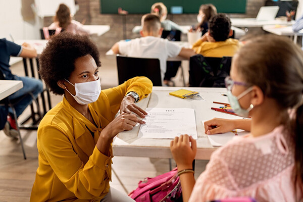 A masked teacher crouches down at a desk to talk with a masked student at school.