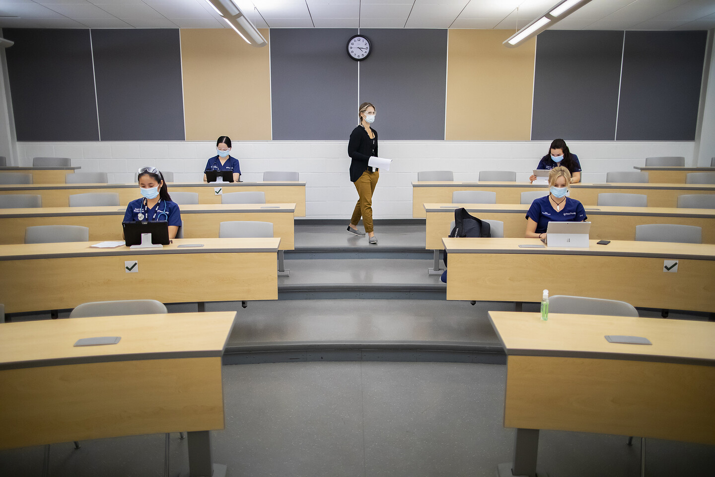 Four students seated at long tables wearing masks, one teacher standing lecturing wearing a mask, in a Penn classroom.