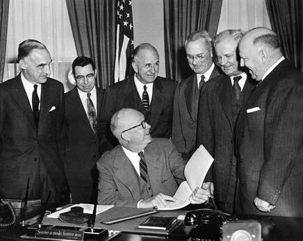 A bald man in glasses and a suit and tie sits at a table pointing to a piece of paper and smiles as he looks to his left, surrounded by six other men behind him