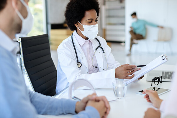 Medical doctor wearing a face mask and stethoscope sits at a desk holding a pen and paper going over details with a masked patient.