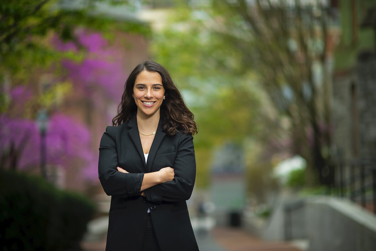 Woman in a black business suit crosses her arms and smiles into the camera, standing on a path with trees in pink spring bloom and new green leaves along a path behind her