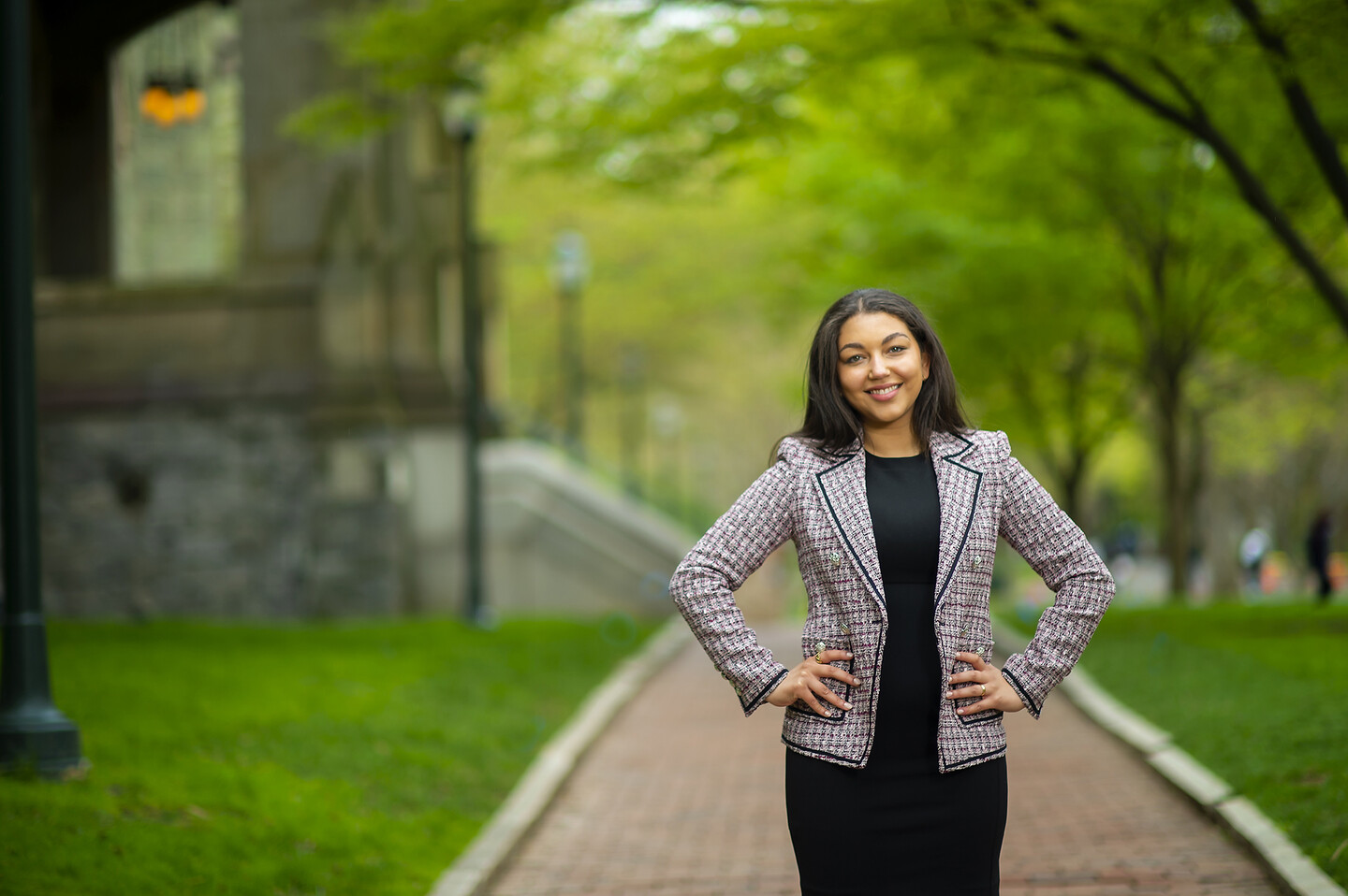 Woman a black dress and a pink, black and white blazer with her hands on her hips stands on a brick path with a building and staircase behind her on the left, green grass on either side of the pathway and green budding trees in the background.