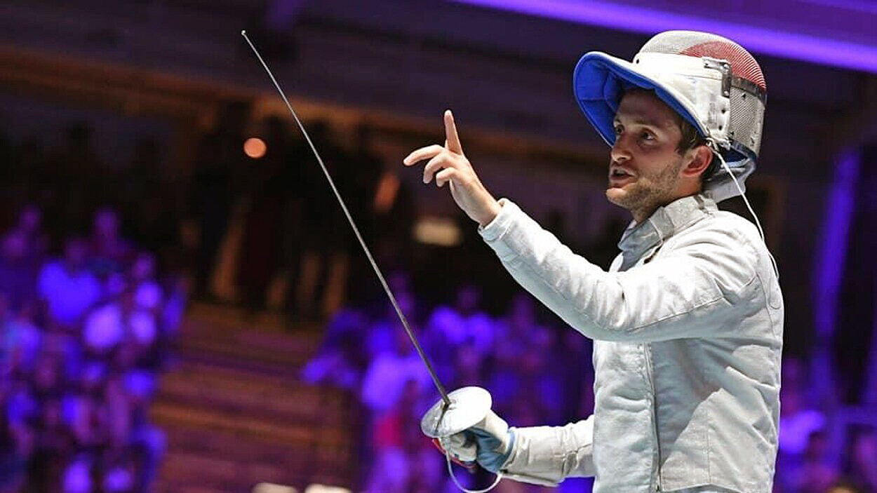 Shaul Gordon in fencing regalia holds his hand up while clutching a fencing sabre in the other.