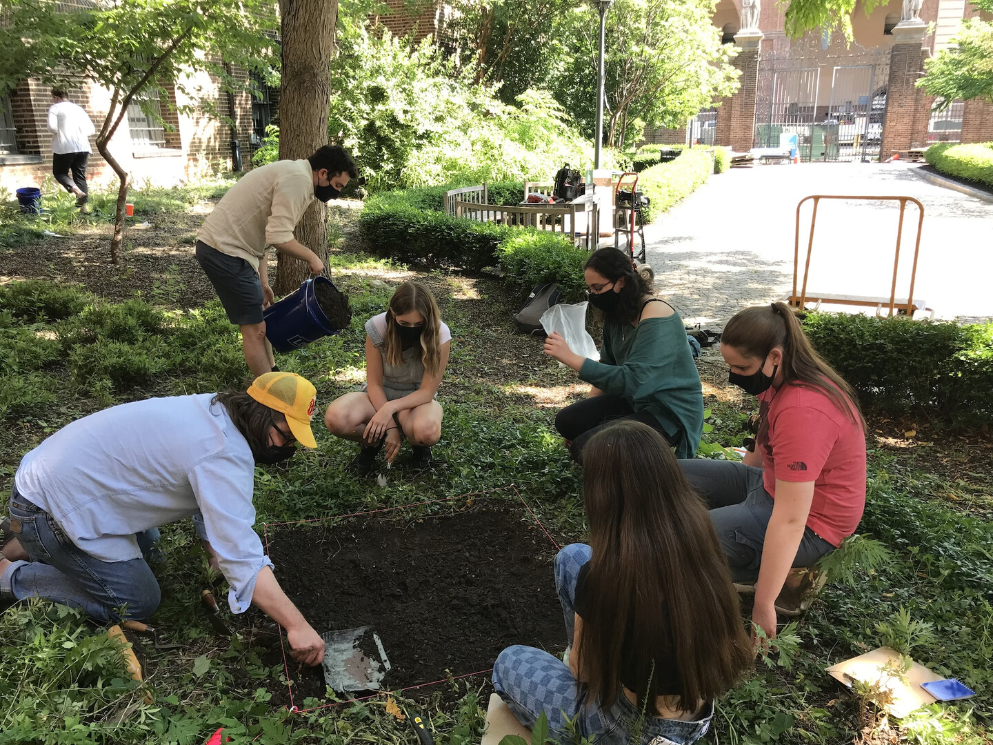 A group of people surrounding a roped-off patch of dirt in a courtyard area. One is using a dustpan to smooth the dirt. Another is dumping a 5-gallon bucket of mulch. Several others are on the ground looking at the site.