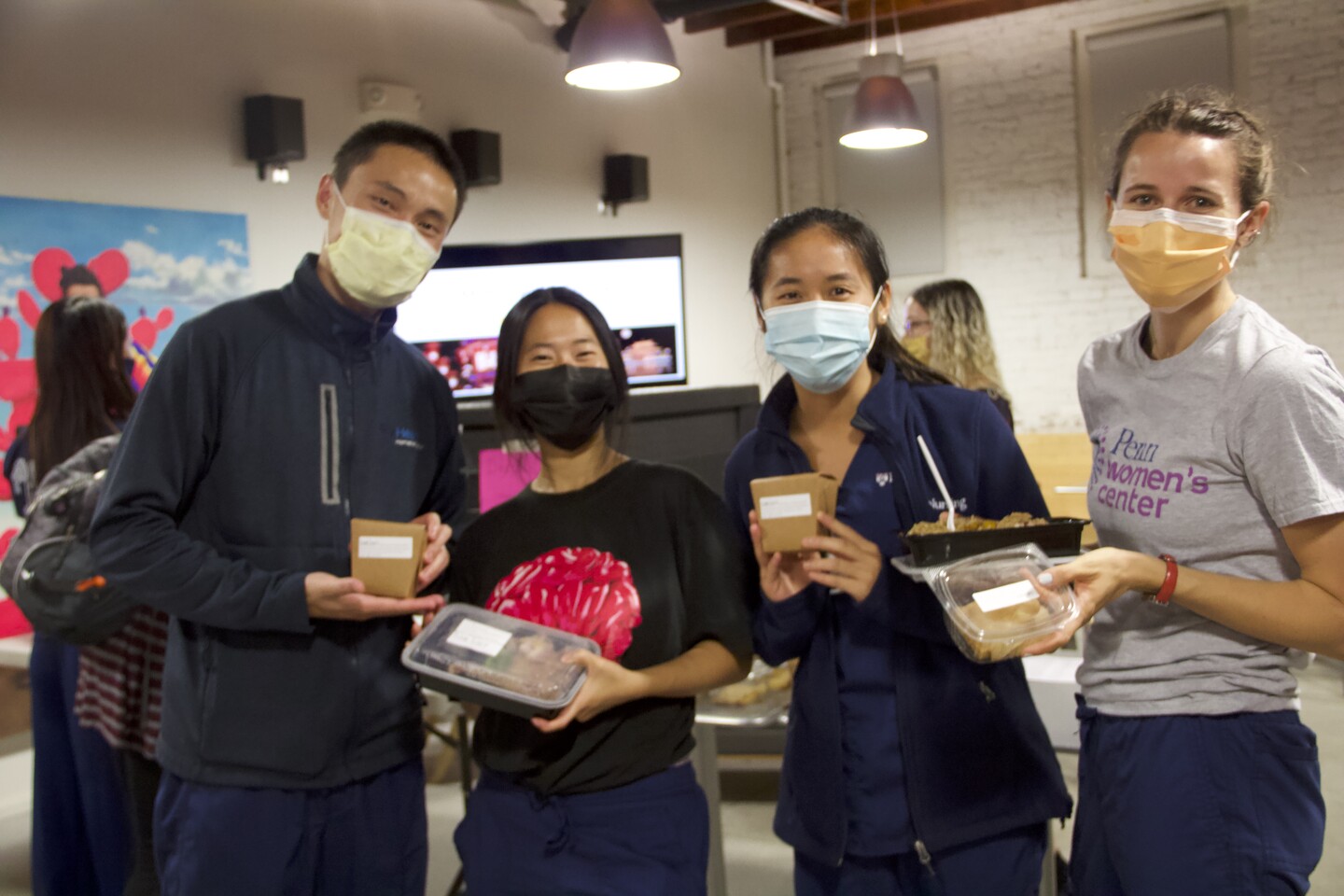 Four students wearing blue hold up their boxed dinners. One student wears a shirt saying "Penn Women's Center"