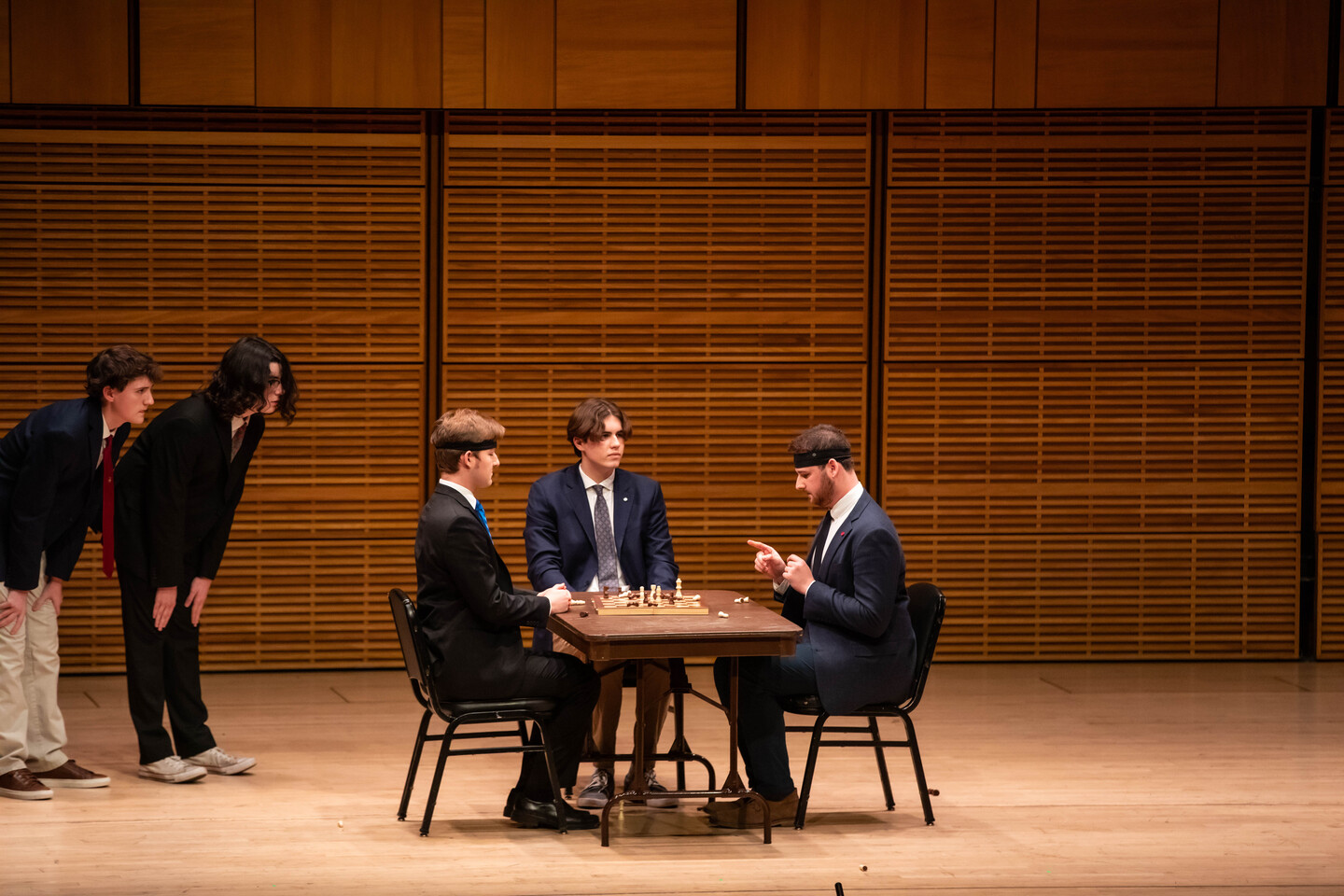 three students sitting at a table and two standing nearby on a stage