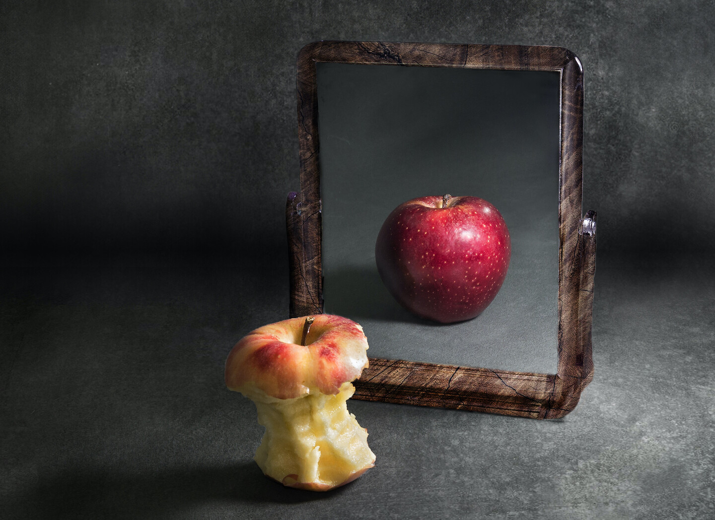 A mostly eaten apple in front of a mirror showing a whole, uneaten apple. 