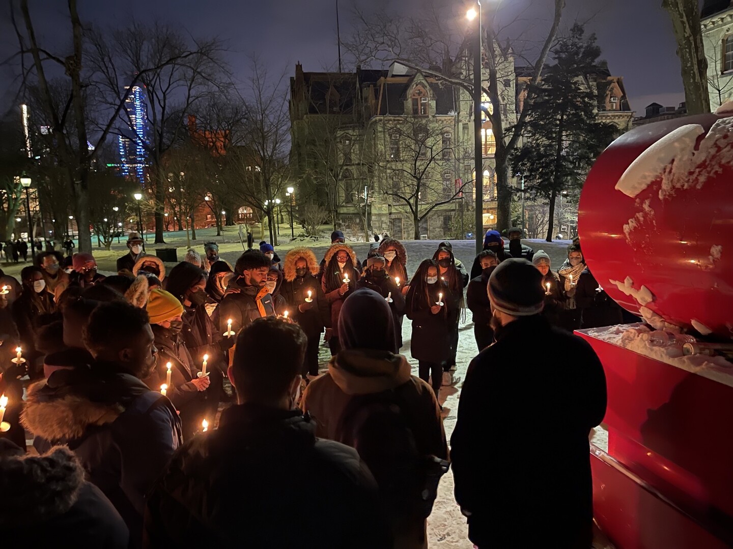 A circle of people stand holding candles in the snow beside the "LOVE" statue