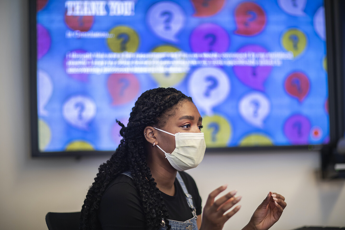 Adetobi Moses sitting in front of a blurry large screen, wearing a mask. 