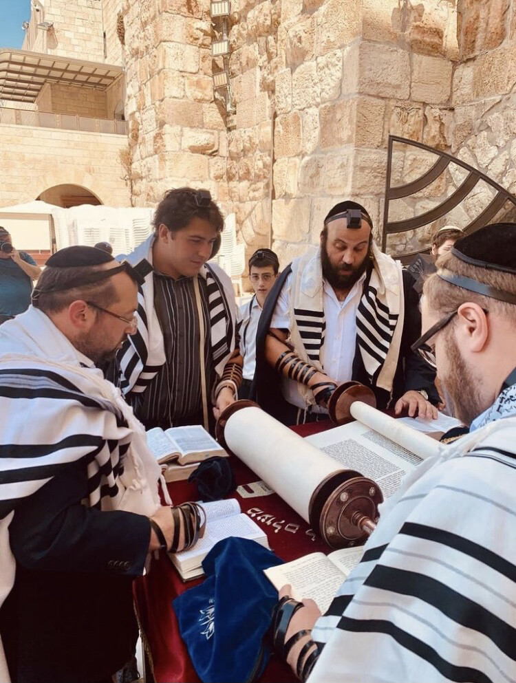 Cam Landis reads from a scroll rolled out on a table. Three men surround him, all wearing prayer scarves.’